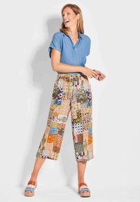 Denim polo shirt met opgerolde mouwen, gecombineerd met patchwork broeken in verschillende kleuren en patronen, en chunky sandalen met meerkleurige bandjes.