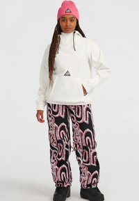 Pull-over blanc avec capuche et poche avant, associé à un pantalon noir avec un motif abstrait rouge et blanc. Bottes noires et bonnet rose.