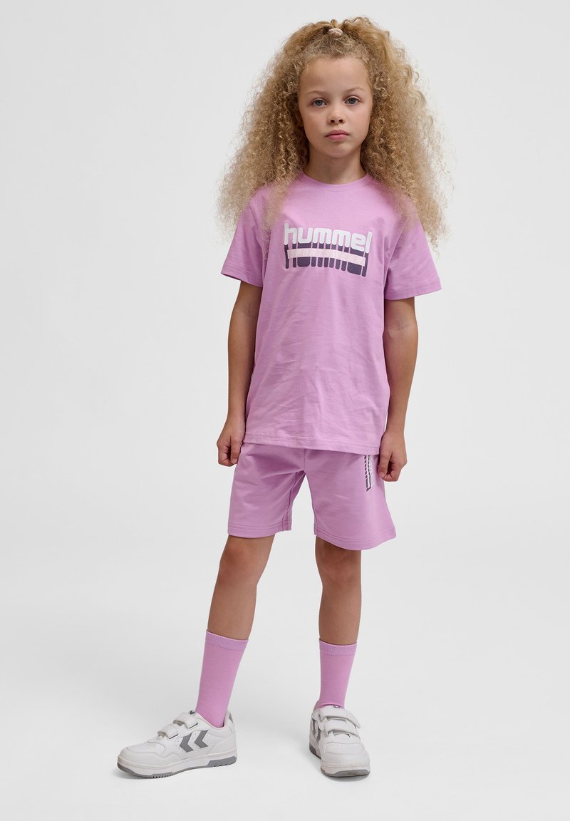 Lilla T-shirt med "hummel" logo, parret med matchende shorts og hvide sneakers. Har krøllet hår og lyserøde sokker.