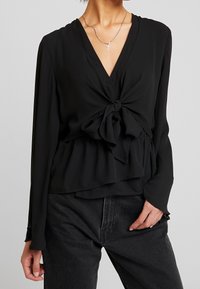 Černá halenka s hlubokým výstřihem do V a uzlem vpředu. Dlouhé rukávy a vrstvený peplum střih. Hladká, lehká tkanina.