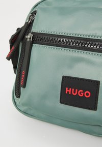 Borsa a tracolla verde in nylon con una cerniera nera, etichetta logo rossa e doppie cerniere con tiranti neri. Superficie testurizzata con dettagli di cucitura.