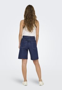 Shorts en denim de lavage foncé, longueur mi-cuisse, avec un design classique à cinq poches et des coutures contrastées. Portés avec un débardeur blanc.