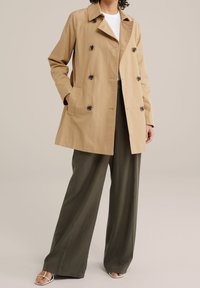 Beżowy trencz z dwurzędowym zapięciem, czarnymi guzikami i bocznymi kieszeniami. Połączony z oliwkowozielonymi spodniami o szerokich nogawkach i sandałami.