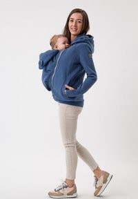 Felpa in pile blu con zip, tasche frontali e inserto per neonati attaccato. Pantaloni chiari e sneakers multicolori.