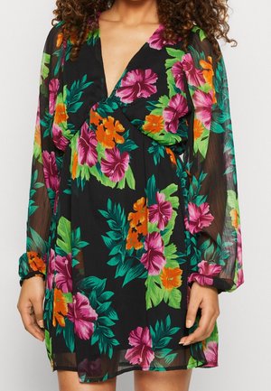 Frau trägt ein schwarzes, transparentes Kleid mit langen Ärmeln und lebendigem pinkem, orangefarbenem und grünem Blumenmuster, mit tiefem V-Ausschnitt.