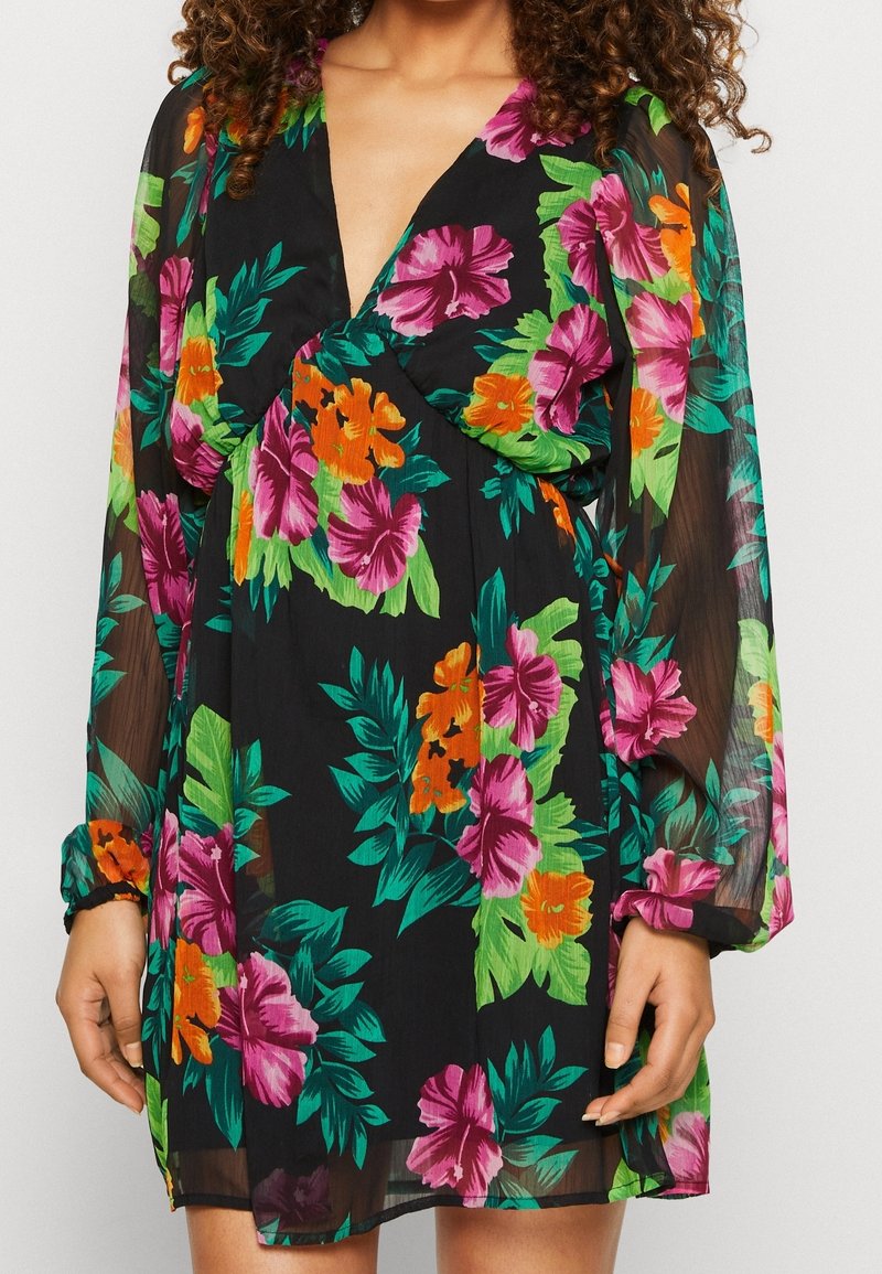 Femme portant une robe noire transparente à manches longues avec un imprimé floral vif rose, orange et vert, avec un décolleté en V profond.