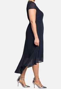 Robe mi-longue bleu marine avec des manches assorties transparentes et une jupe fluide. Associée à des chaussures à talons hauts argentées brillantes avec des brides à la cheville.