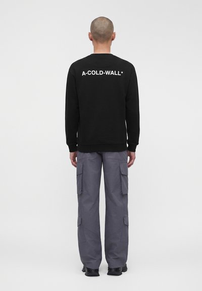 Άνδρας με ξυρισμένο κεφάλι, γυρισμένος με την πλάτη, φορά μαύρο φούτερ με το κείμενο «A-COLD-WALL*» και γκρι παντελόνι cargo με μαύρα παπούτσια.