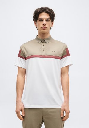 Junger Mann mit dunklem lockigem Haar, der ein Polohemd in Beige, Weiß und Weinrot trägt und vor einem schlichten weißen Hintergrund steht.