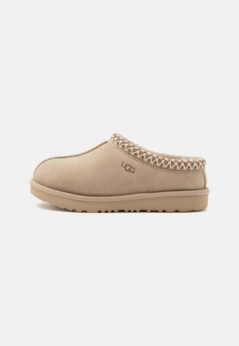 Chausson beige en daim à enfiler avec logo UGG, couture décorative autour de l'ouverture et semelle épaisse en caoutchouc pour usage intérieur et extérieur.
