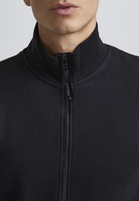 Veste noire à zip avec col montant, fabriquée en tissu doux. Présente une fermeture éclair centrale et un design minimaliste.