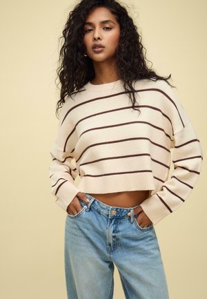 Pull court crème avec des rayures marron foncé ; poignets et ourlet côtelés. Jean taille haute bleu clair avec une coupe décontractée.