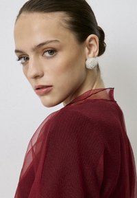 Pendientes de botón de cristal transparente combinados con una blusa burdeos translúcida con un tejido texturizado, que muestra un escote suave y elegante.