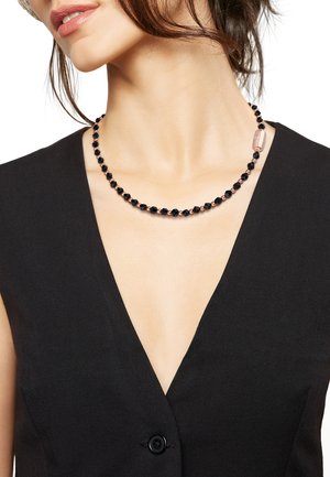 Collana di perline nere con perline sfaccettate nere e un accento metallico rosa in un design semplice, indossata sopra un top nero a V.