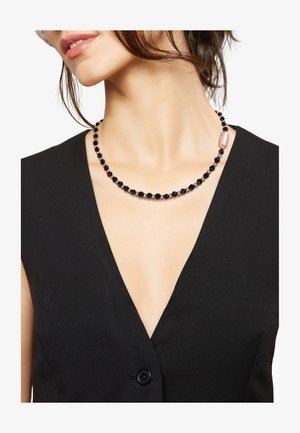 Schwarze Perlenkette mit facettierten schwarzen Perlen und einem metallischen pinken Akzent in einem schlichten Design, getragen über einem schwarzen V-Ausschnitt-Top.