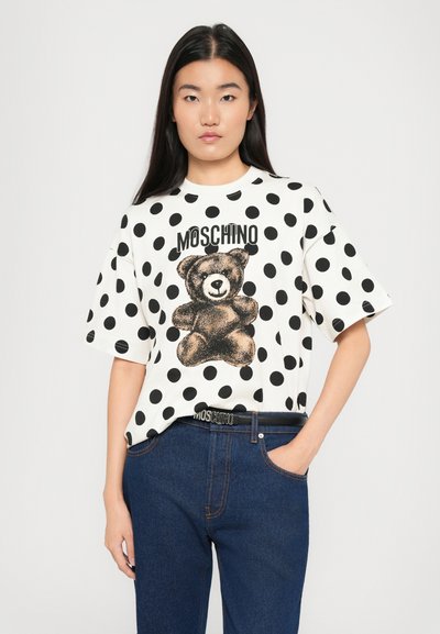 Noor naine pikal tumedal juuksel, seljas valge täpiline t-särk, millel on kaisukaru ja "Moschino" tekst, koos siniste teksade ja Moschino vöödiga.