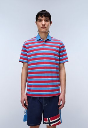 Mand iført en rød og lyseblå stribet polo-shirt og marineblå shorts med en rød, hvid og blå flagpatch, står mod en ensfarvet lys baggrund.