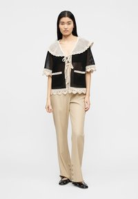 Γυναίκα που φοράει μαύρο τοπ από δίχτυ με μπεζ κέντημα κρόσσια και μπεζ παντελόνι φαρδύ με λεπτομέρειες κουμπιών στους αστραγάλους, στέκεται μπροστά σε λευκό φόντο.