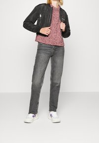 Person in schwarzer Bomberjacke über pinkem Strickpullover, grauen Jeans und weißen Sneakers mit lila Details, der vor einem schlichten Hintergrund steht.