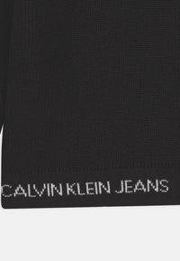 Czarna dzianinowa talia z białym napisem "CALVIN KLEIN JEANS". Teksturowana tkanina o płaskim, dopasowanym kroju. Brak dodatkowych wzorów.