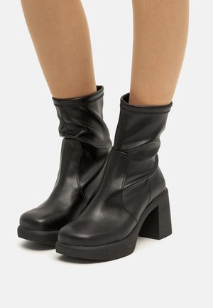 Botas de tobillo de cuero negro con un diseño arrugado, tacones altos en bloque y una suela texturizada. Presentan costuras mínimas y un acabado suave.