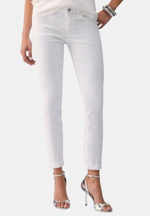 Donna che indossa jeans bianchi aderenti con orli risvoltati e sandali con tacchi a spillo argento, con braccialetti argentati a un polso.