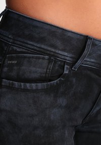 Jeans de denim negro con una textura desgastada, que cuentan con un bolsillo frontal y un sutil logo "RAW" en la cinturilla. Diseño clásico de cinco bolsillos.