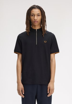 Homme avec de longues dreadlocks portant un polo noir à manches courtes avec une fermeture éclair à l'avant et un petit emblème doré sur la poitrine.