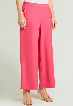 Woman wearing bright pink wide-leg pants and strappy gold high-heeled sandals, standing against a plain light background.