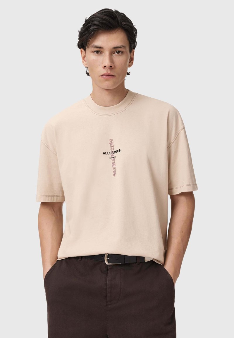Camiseta de algodón beige con un corte relajado, presenta un logotipo negro central y un gráfico de texto. Diseño de mangas cortas y cuello redondo.