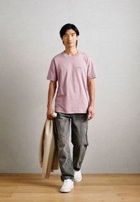 T-shirt rosa chiaro a maniche corte con taschino, abbinata a jeans grigi larghi e sneaker bianche. Tiene una giacca beige sul braccio sinistro.