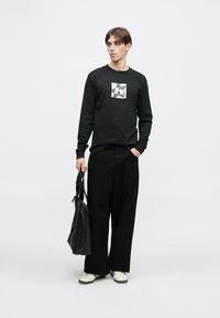 Černé tričko s dlouhým rukávem s grafickým logem, doplněné volnými černými kalhotami a bílými teniskami. Model drží černou tašku přes rameno.