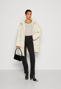 Manteau rembourré crème, haut rayé, jeans noirs, bottes à talons noires et un sac à main structuré. Texture douce, cet vêtement d'extérieur est doté d'une capuche.