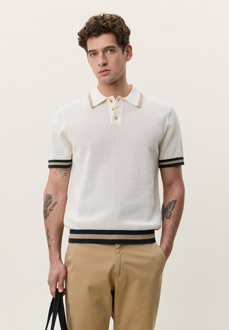 Homme aux cheveux bouclés portant un polo en maille blanc et un pantalon beige, tenant un sac noir avec des tatouages sur les deux avant-bras.