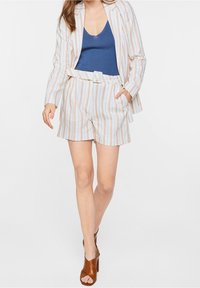 Kvinna som bär en beige och vit randig blazer och shortset med en blå ärmlös topp och bruna öppna klackar, poserar med händerna i fickorna.