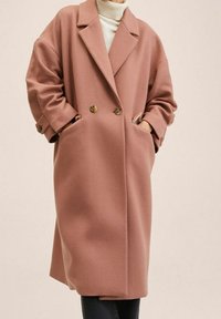 Manteau oversize pour femme en laine rose poussiéreuse avec larges revers, double boutonnage et mains dans les poches, porté sur un pull à col roulé blanc.