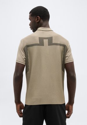 Mand iført beige kortærmet skjorte med sort lineært korsmønster på ryggen, kombineret med sorte bukser, vendt væk mod hvid baggrund.