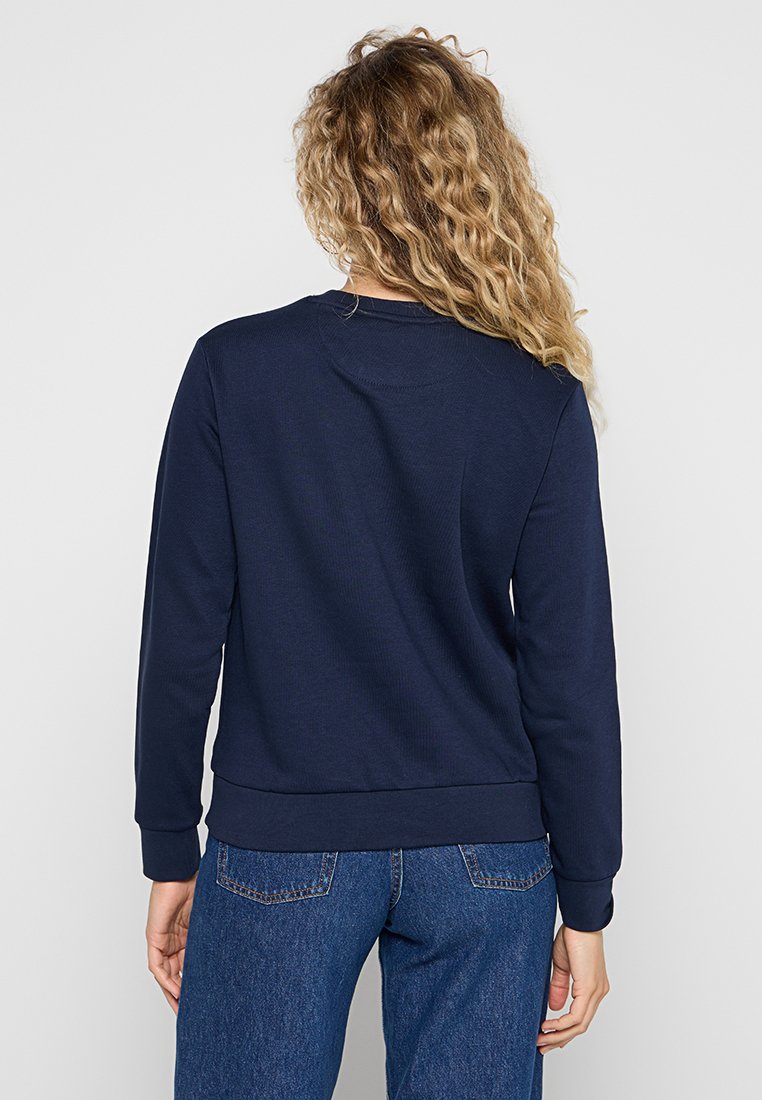 Frau mit lockigem blonden Haar, die ein dunkelblaues Langarmshirt und eine blaue Jeans trägt, von hinten gegen einen schlichten Hintergrund betrachtet.