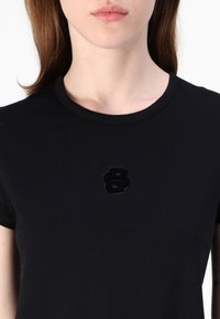 Svart kortermet t-skjorte laget av mykt stoff med et fluffy svart tall "8" brodert på brystet. Rund halslinning, enkel design.