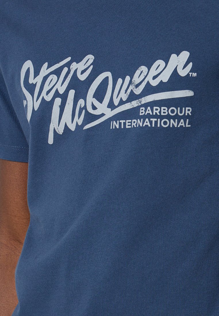 Camiseta de algodón azul con mangas cortas que presenta un estampado gráfico blanco de "Steve McQueen" y "Barbour International". Textura suave.