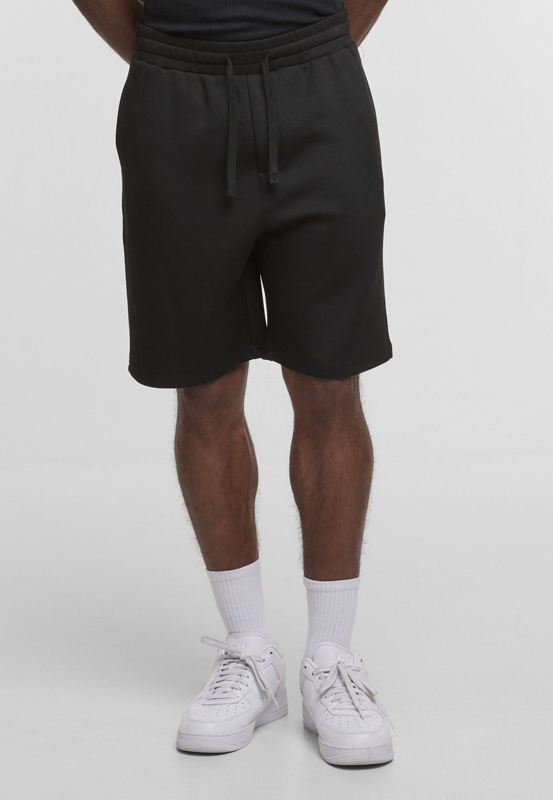 Shorts noirs longueur genou avec cordon de serrage portés par une personne visible de la taille aux pieds, assortis à des chaussettes blanches montantes et des baskets blanches.