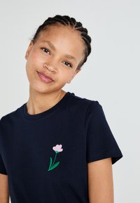 Marineblå bomulls-T-skjorte med en liten rosa blomst brodert på venstre bryst, med grønne blader. Korte ermer og rund hals-design.