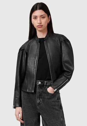 Giacca di pelle nera con design corto e spalle bombate. Presenta zip in argento e un colletto minimalista, abbinata a jeans scuri.