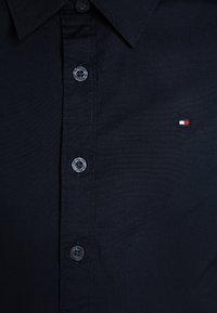 Camisa negra abotonada con cuello puntiagudo, con un logo cerca del pecho y botones redondos negros con el nombre de la marca grabado.