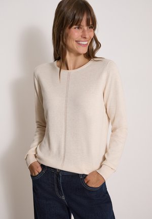 Kvinde smiler og har en cremefarvet langærmet sweater og mørkeblå jeans på med hænderne i lommerne mod en ensfarvet baggrund.