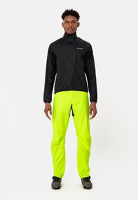 Giacca nera leggera con colletto alto e tasca con zip, abbinata a pantaloni impermeabili verde neon e calzature nere robuste.