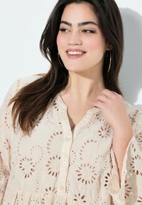 Helle-beige Bluse mit floralem Stickerei-Ausschnitt, einer geknöpften Vorderseite, langen Ärmeln mit ausgestellten Abschlüssen und zartem Textilstoff.