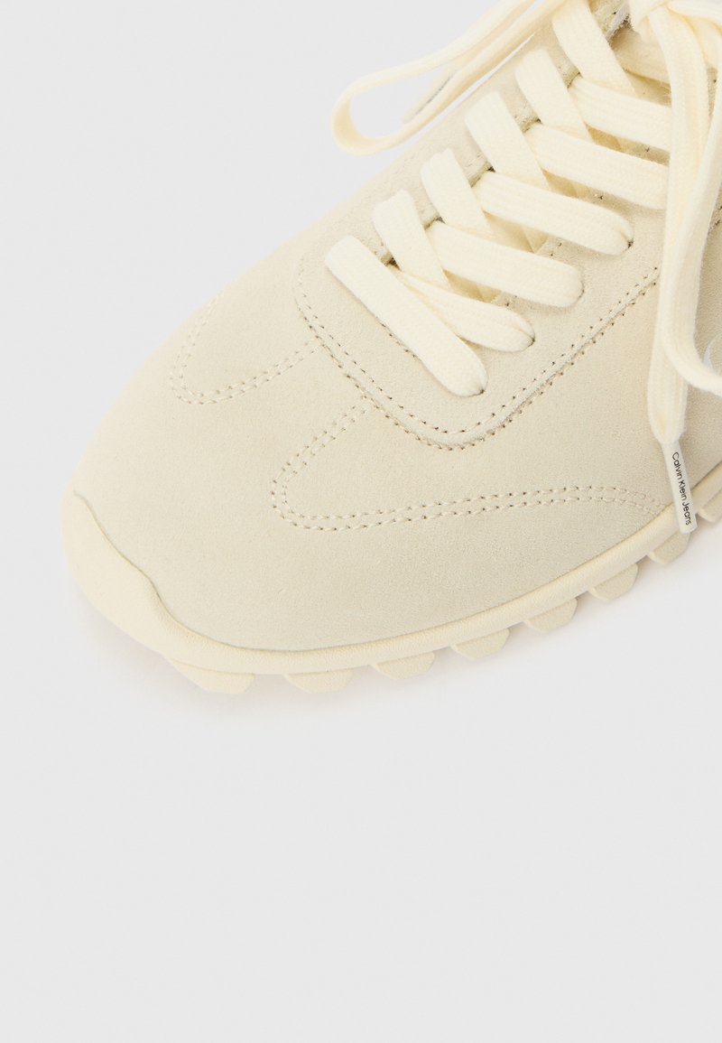 Zapatilla de ante beige claro con cordones planos, puntera redonda y suela de goma texturizada. Presenta costuras decorativas y una marca sutil.