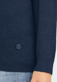 Mörkblå stickad tröja med texturerad yta, med en cirkulär svart logoknapp och ribbade mudd och nederkant.