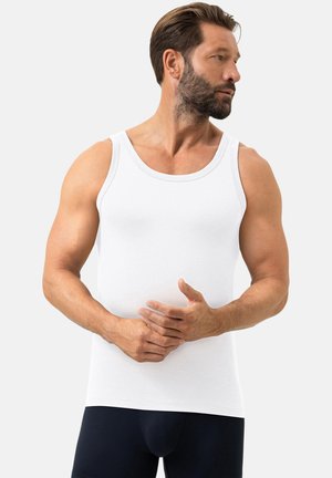 Mann mit Bart, der ein weißes Tanktop und dunkle Hosen trägt, steht und schaut nach rechts, die Hände vor dem Körper verschränkt.